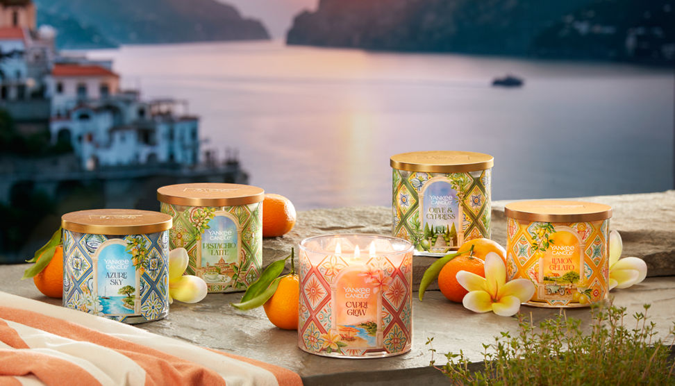 Hello, Italy! Collection Azure Sky 3-Wick Candle, Pistachio Latte 3-Wick Candle, Capri Glow 3-Wick Candle, Olive & Cypress 3-Wick Candle, and Lemon Gelato 3-Wick Candle on a rock surface surrounded by fruit and flowers overlooking water and a sunset
