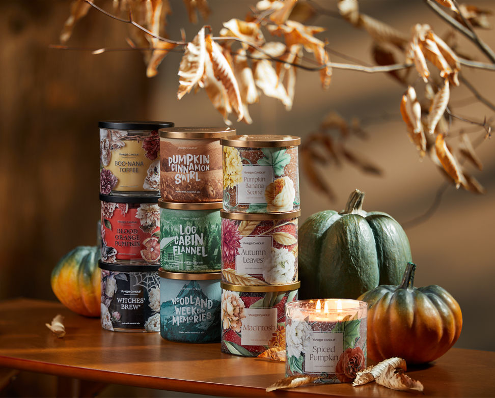 Boo-nana Toffee 3-Wick Candle, Blood Orange Pumpkin 3-Wick Candle, Witches' Brew® 3-Wick Candle, Pumpkin Cinnamon Swirl 3-Wick Candle, Log Cabin Flannel 3-Wick Candle, Woodland Weekend Memories 3-Wick Candle, Pumpkin Banana Scone 3-Wick Candle, Autumn Leaves® 3-Wick Candle, Macintosh 3-Wick Candle, and Spiced Pumpkin 3-Wick Candle on a wood table with decorative pumpkins and leaves
