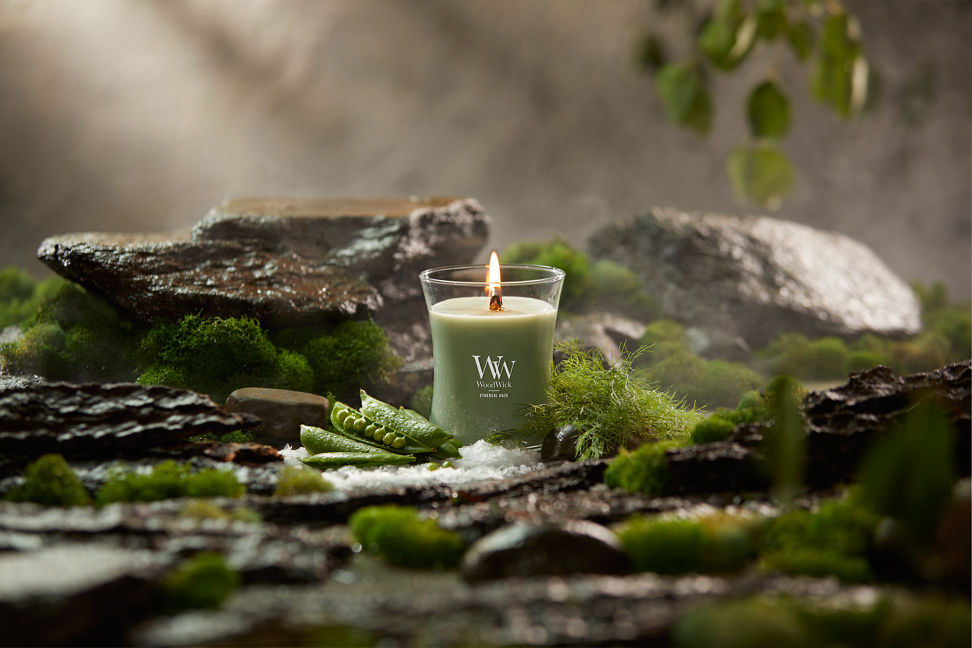 WoodWick Vapour Trails Collection Ethereal Haze Medium Hourglass Candle surrounded by moss, rocks, salt, and snap peas in a forest setting