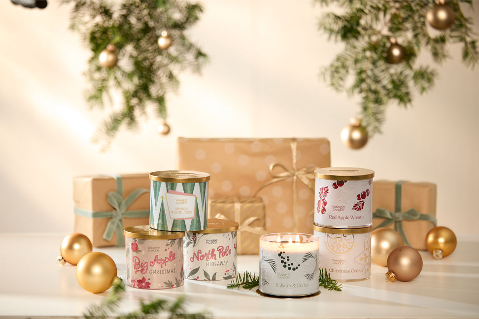 Magical Frosted Forest 3-Wick Candle, Balsam & Cedar 3-Wick Candle, Red Apple Wreath 3-Wick Candle, and Christmas Cookie 3-Wick Candle with Passport to the Holidays Collection Big Apple Christmas 3-Wick Candle and North Pole Hideaway 3-Wick Candle on a table next to wrapped gifts and gold ornaments