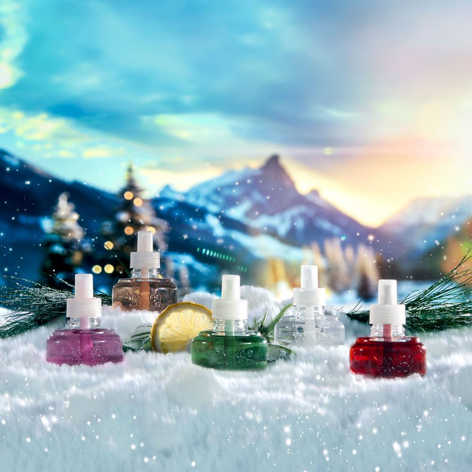 an array of apres ski scentplug refills in the snow with pine needles and herbs and sliced lemon with a mountain scene behind