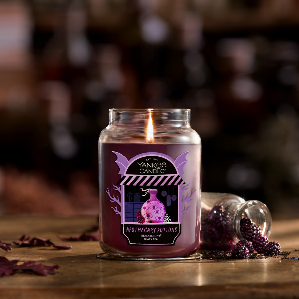 Large purple jar candle with apothecary label lit on rustic table beside spilled decorative blackberries