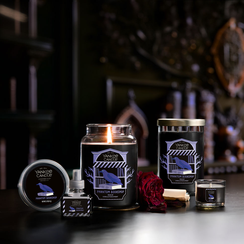 Large black jar candle with raven label, surrounded by rose, books, and other candles in a cozy home setting