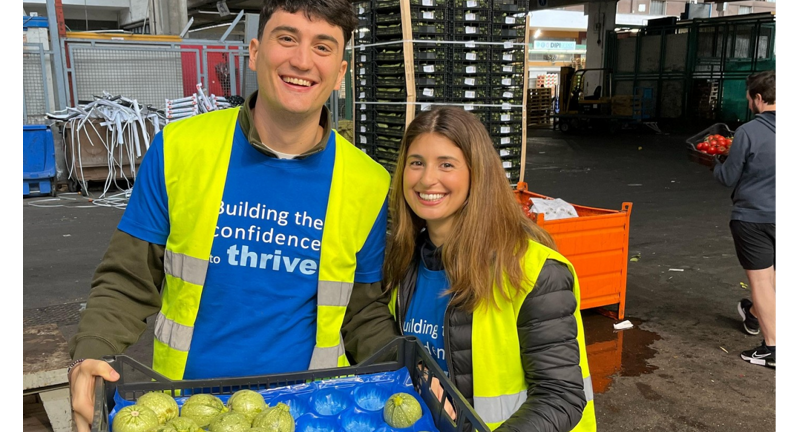 Two individuals with bright yellow vests are shown holding a tray of vegetables in a food distribution center, exemplifying volunteer work supporting food security.
