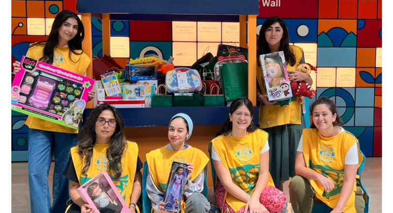 A group of children, alongside adults, is standing in a brightly colored area holding toys and games, participating in a charity event to distribute gifts.