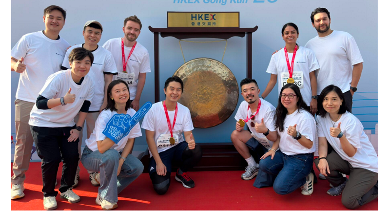 A group of people is shown posing together in matching white t-shirts after completing a charity run event, celebrating their participation with medals in front of a large gong.