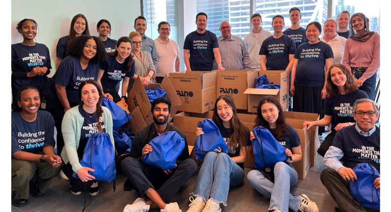 A group of volunteers stands in an office space, surrounded by boxes and bags, all wearing matching blue shirts that say “Building the confidence to thrive”, working on community service projects.