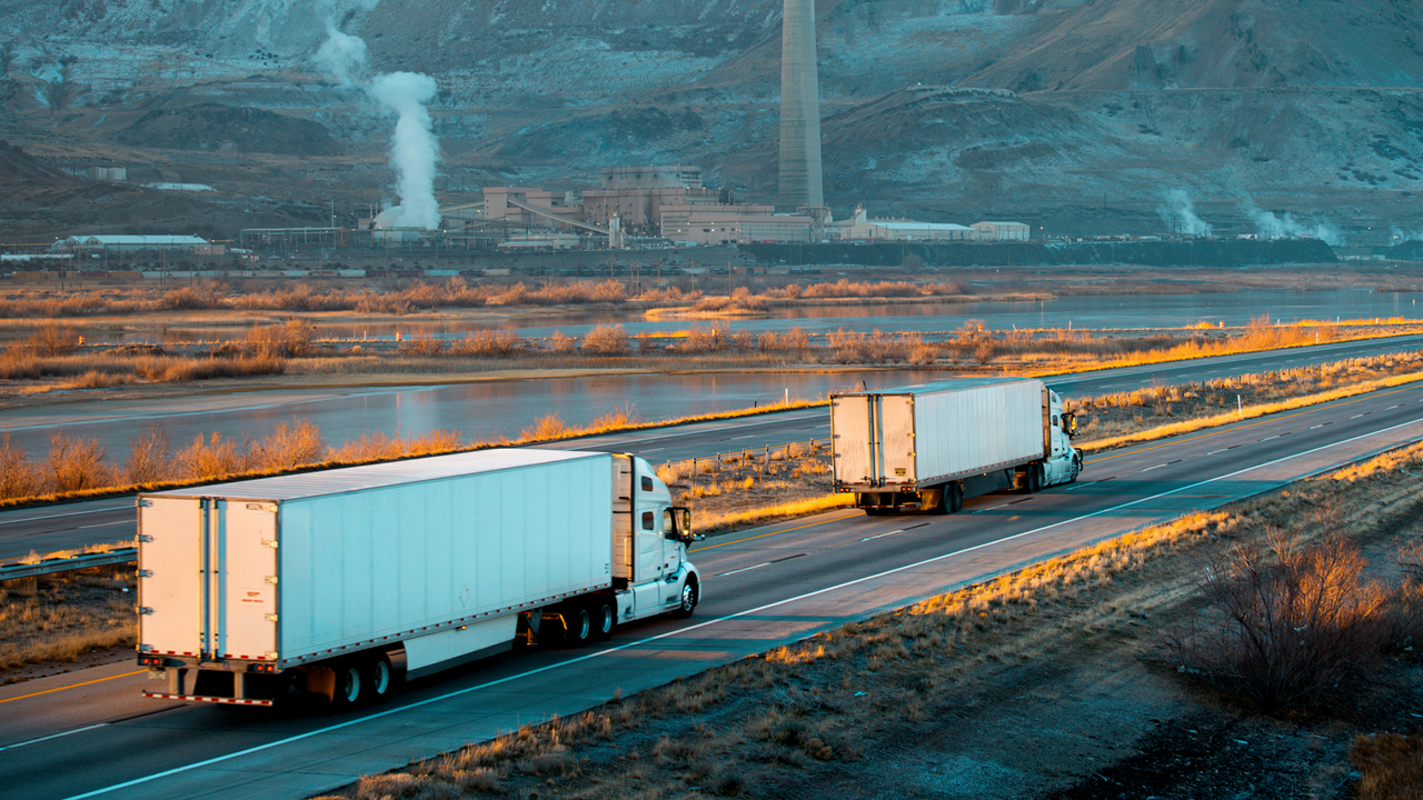 Two semi-trucks travel along a highway beside an industrial plant, with mountains and steam rising in the background.