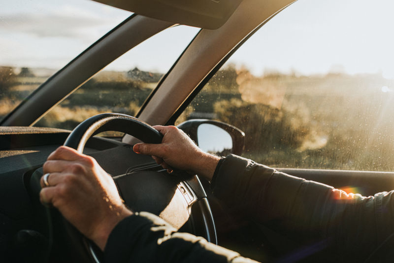 Interior of a car. Hand holds a black steering wheel. Car is illuminated by low, warm sun at dusk.Link: https://www.gettyimages.com/detail/photo/Hand-holding-steering-wheel-in-a-Car-royalty-free-image/1294306347