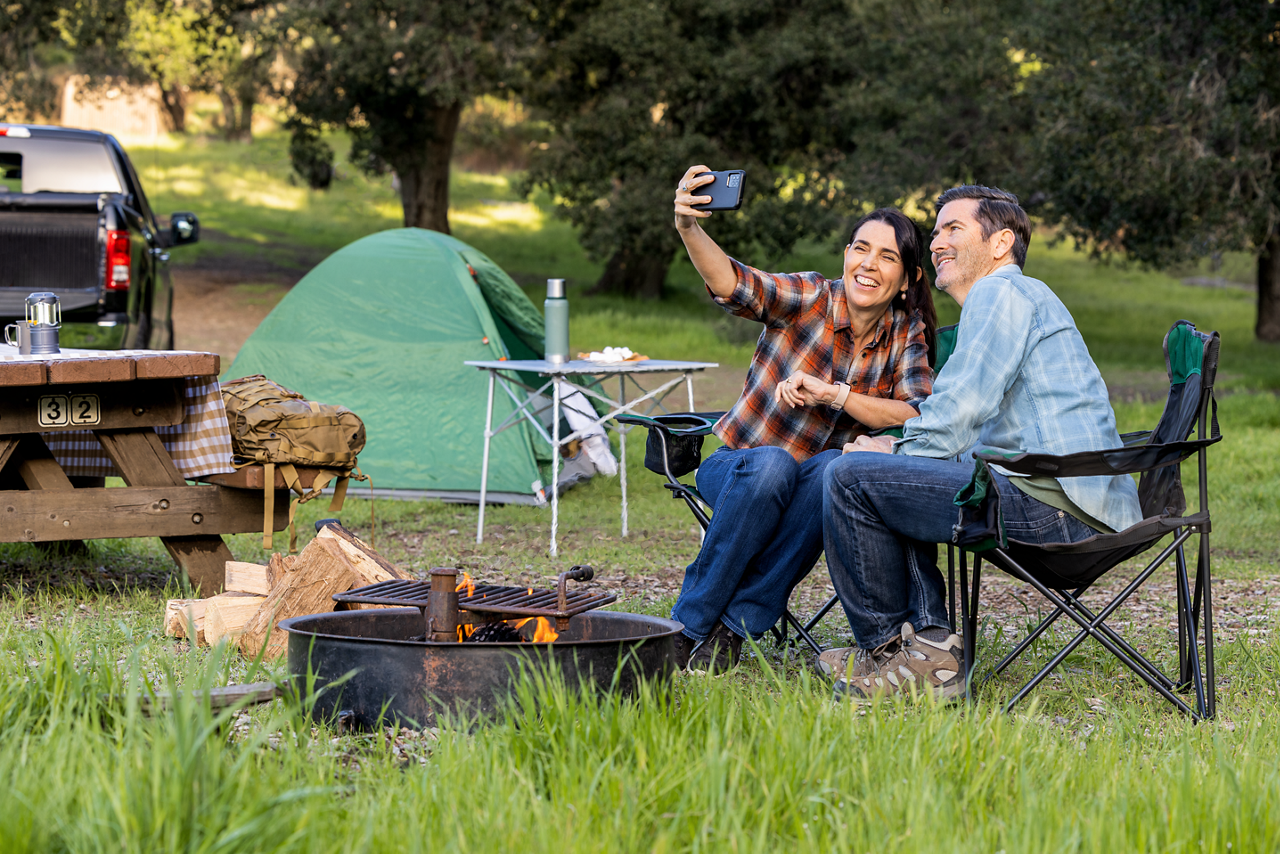 A couple relaxes by a campfire and takes a selfie while camping, with a tent, truck, and forest backdrop. The image represents freedom, adventure, and wireless connectivity in remote settings — perfect for showcasing T-Mobile’s network coverage and lifestyle branding.