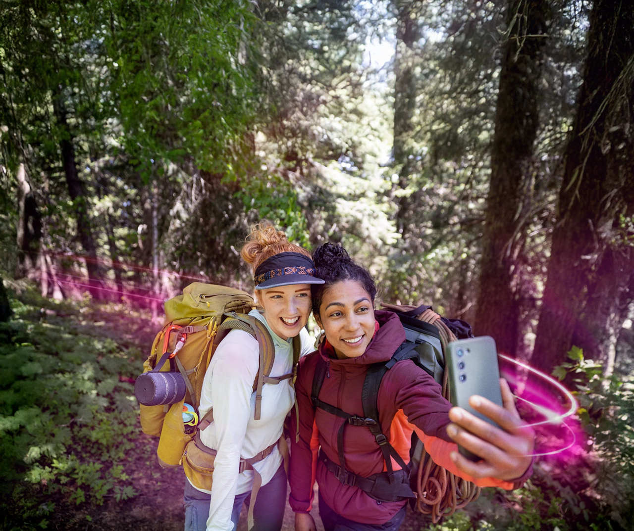 Among tall trees in a forest clearing, two women wearing hiking backpacks and gear smile at a mobile device as magenta beams of light circle them.