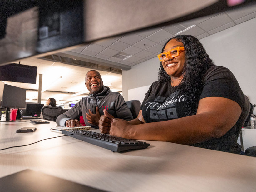 T-Mobile employee smiling while working on a laptop in an office with teammates in the background.