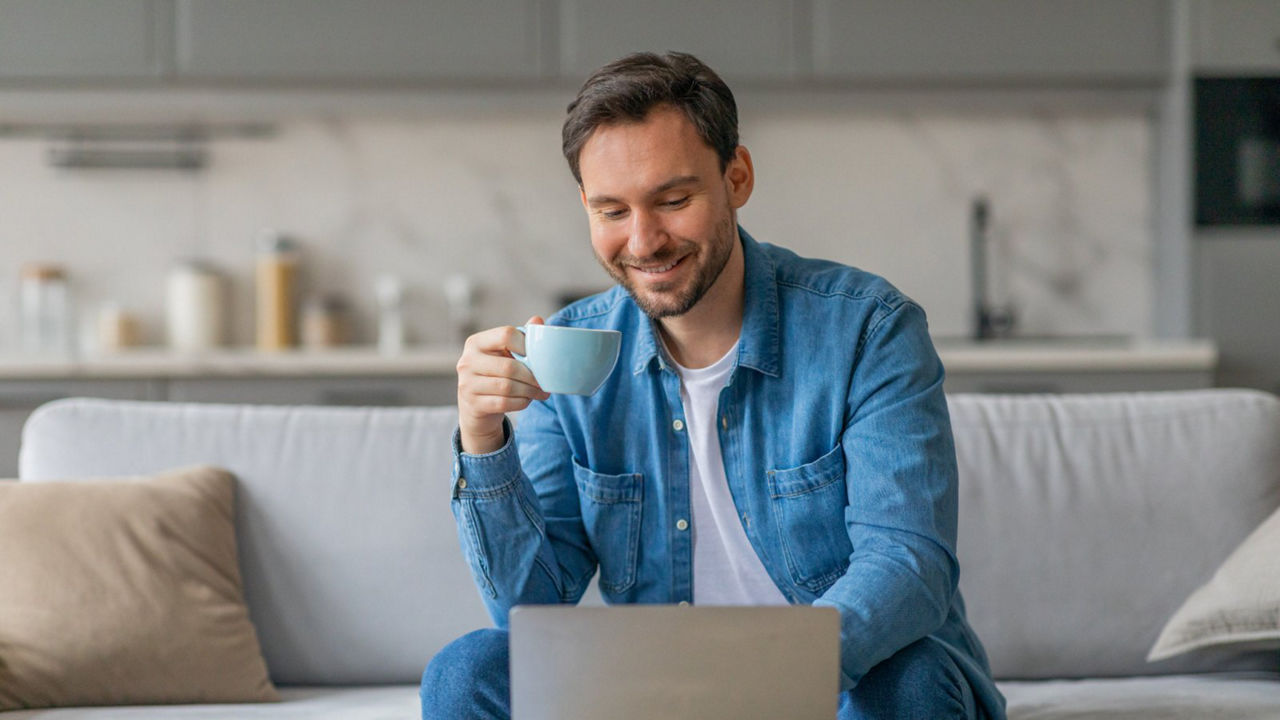 Person sitting on a coach at home holding a cup of coffee while using their laptop