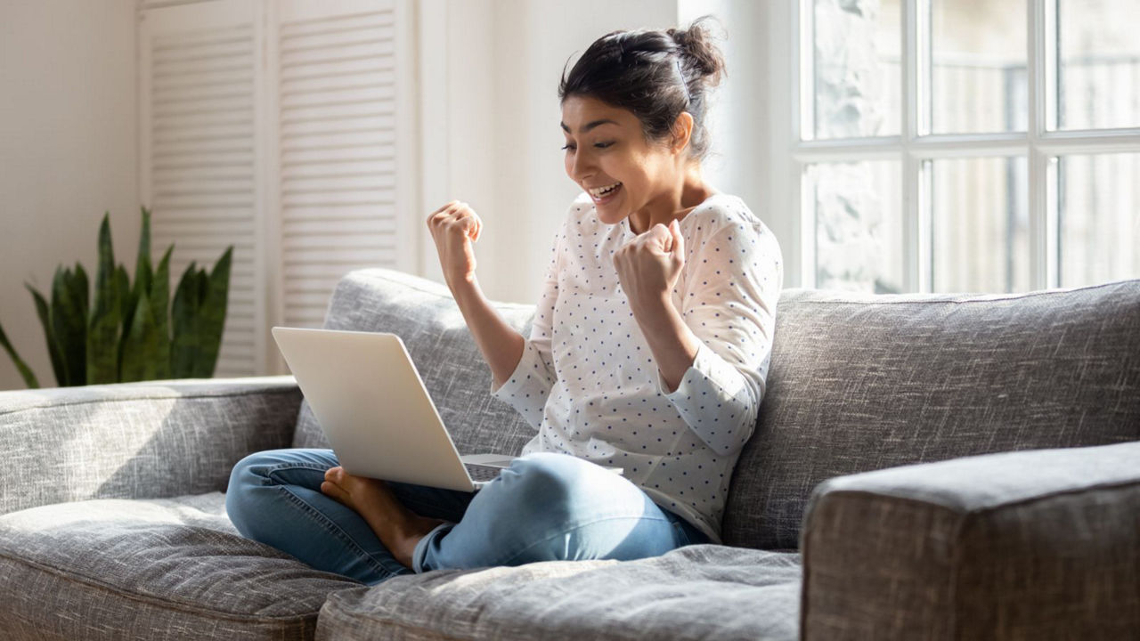 Individual sitting on the couch with legs crossed looking at their laptop celebrating
