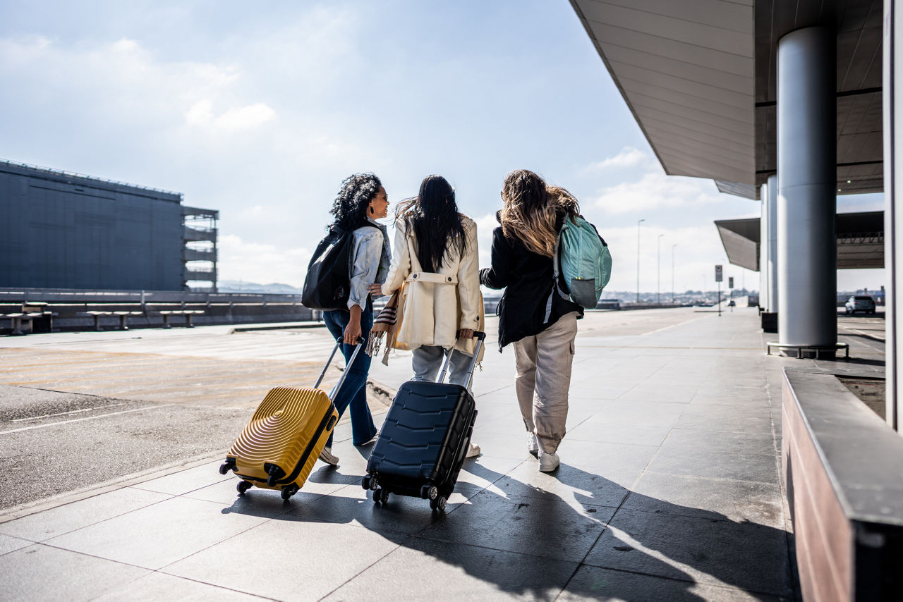 Rear view of three women walking together outside an airport terminal, two pulling rolling suitcases and one carrying a backpack.