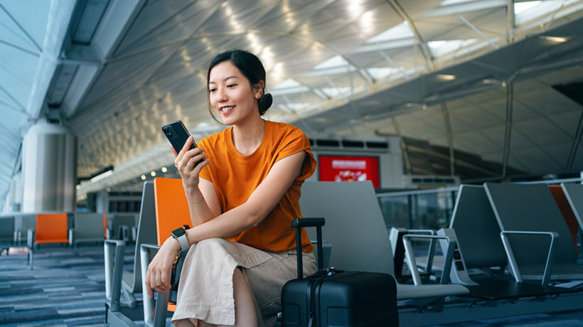 A young woman in an orange t-shirt smiles while using her smartphone at an airport gate, with a black rolling suitcase beside her and rows of empty seats in the background.