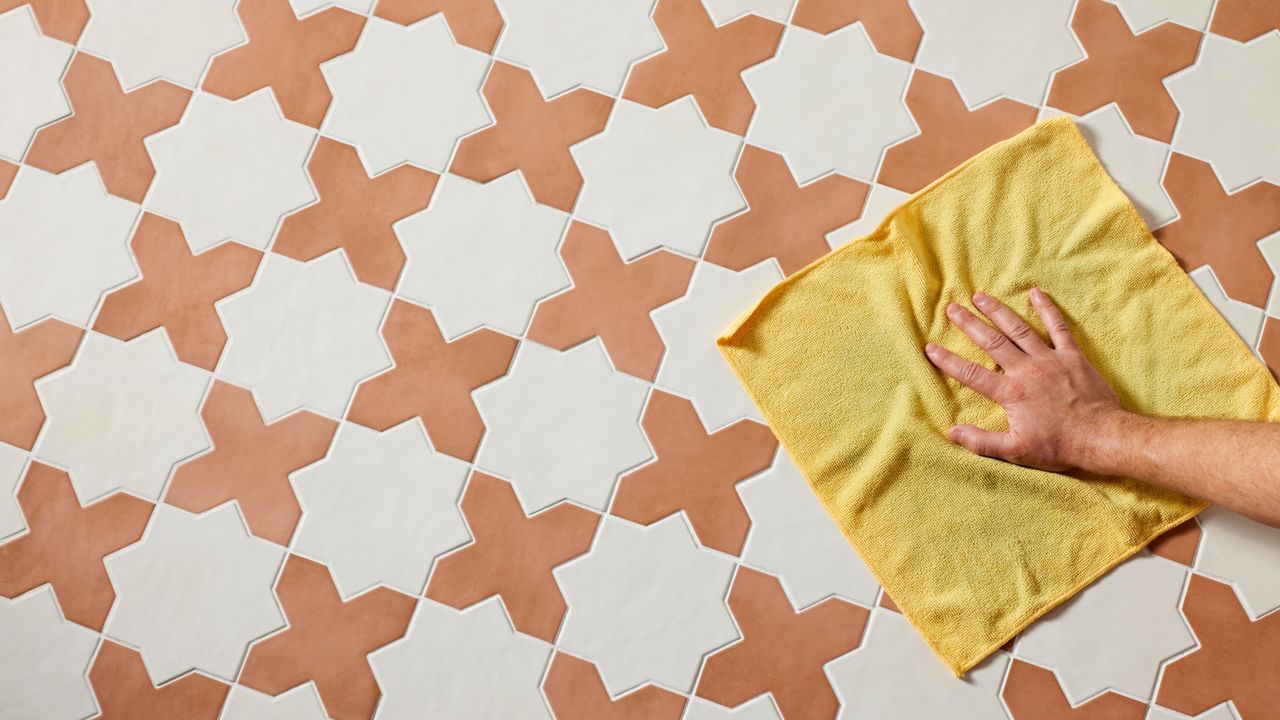 A high-angle view of a person's hand wiping a terracotta and white geometric tile floor with a yellow sponge