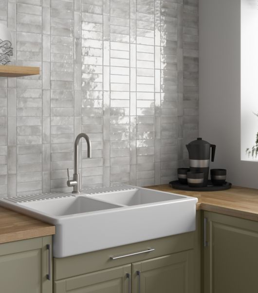 Grey glossy porcelain subway tiles behind a white farmhouse sink, green cabinets and a butcher block counter top.