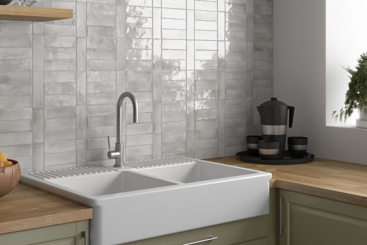 Grey glossy porcelain subway tiles behind a white farmhouse sink, green cabinets and a butcherblock counter top.