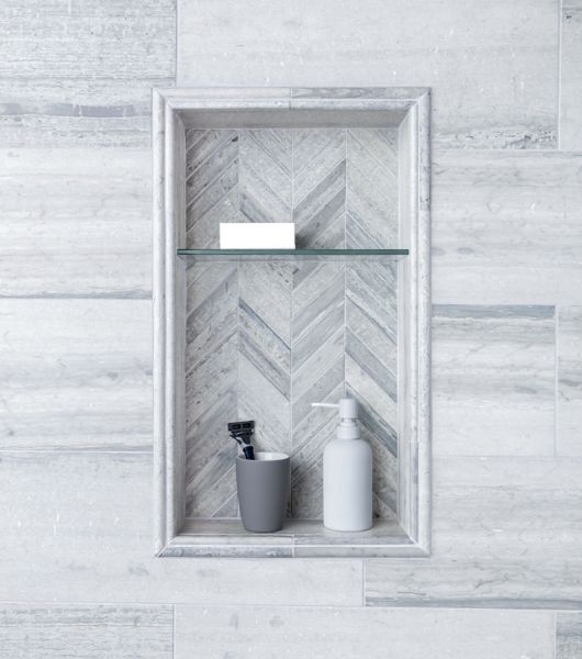 This recessed shower niche in gray tones features a chevron travertine mosaic tile at the back and a single glass shelf. It is framed by coordinating pieces of travertine trim. The main wall of the shower is tiled with a light gray honed travertine subway tile with subtle color variations.