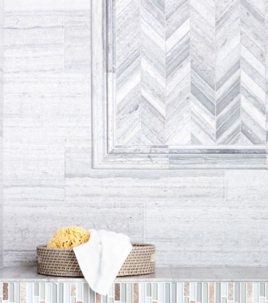 This bathroom features multiple types of limestone tile. A chevron-pattern mosaic is used for an accent wall in the shower, where it is framed with honed travertine trim. The rest of the wall is covered in extra-long travertine subway tiles in shades of cool gray.