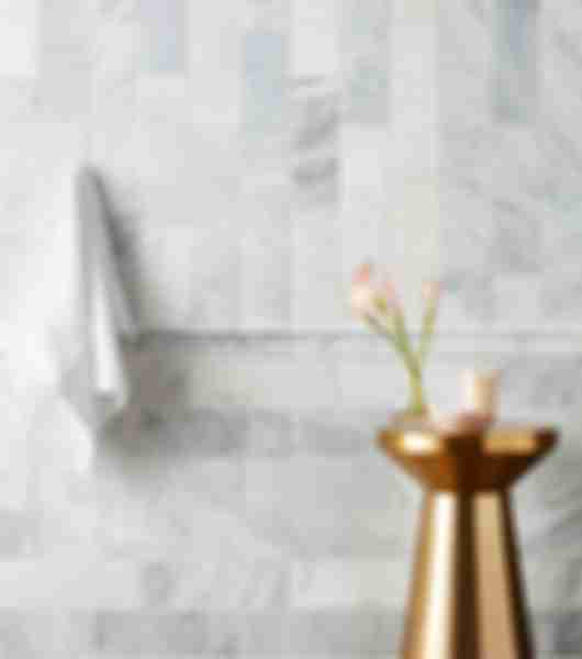 A bathroom wall is covered from floor to ceiling in antique white brushed marble subway tile with gray speckling and tan veining. The tile is arranged in a horizontal off-set pattern from the floor to about halfway up the wall, where a matching trim piece of carved marble extends for the width of the wall, emulating the look of a wooden chair rail. Above this line, the tile is arranged in a vertical off-set pattern, creating contrasting perpendicular lines where the upper and lower halves of the wall meet.