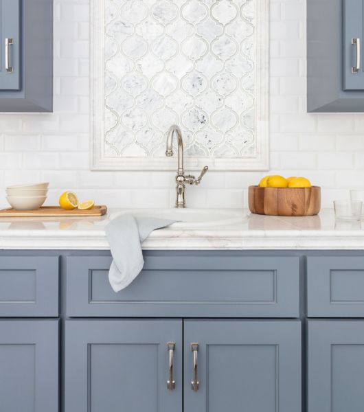 This kitchen features upper and lower cabinets in a medium blue-gray color, white marble countertops, and an over-sink accent wall that uses stone-and-glass mosaic tile against a background of beveled white ceramic subway tile.