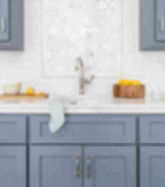 This kitchen features upper and lower cabinets in a medium blue-gray color, white marble countertops, and an over-sink accent wall that uses stone-and-glass mosaic tile against a background of beveled white ceramic subway tile.