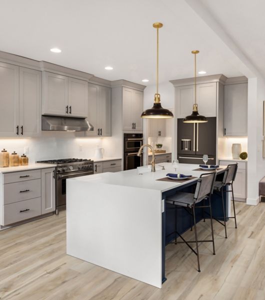 This large open-plan kitchen features a large white waterfall island and light gray upper and lower cabinetry. The floor is covered in long planks of beige wood-look luxury vinyl tile.