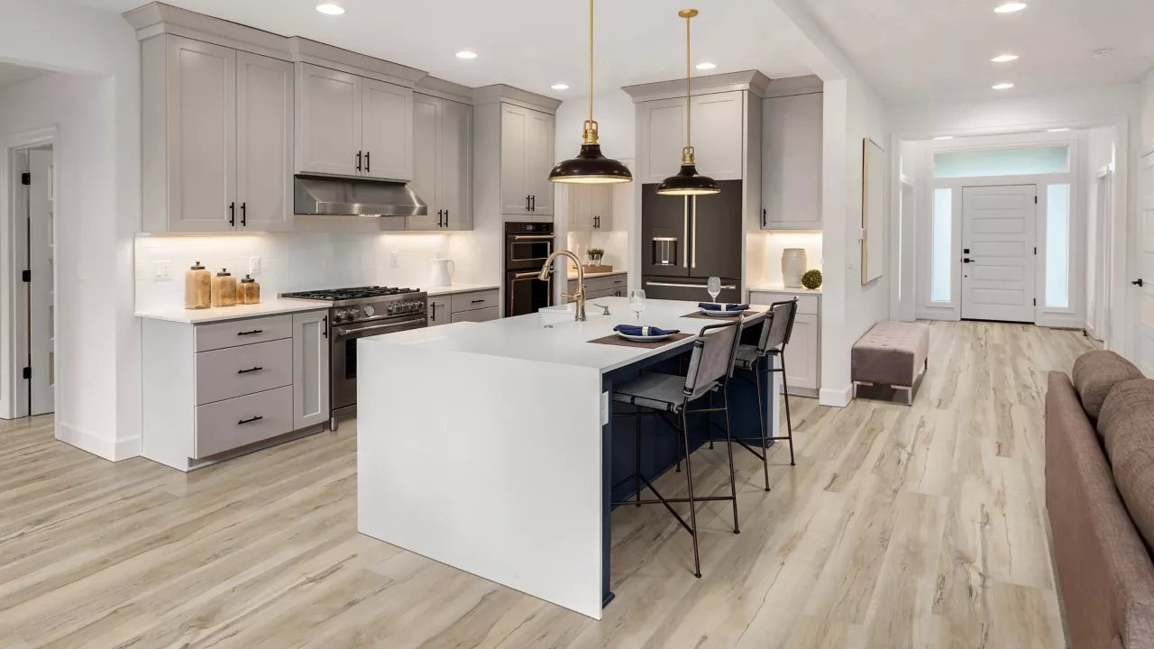 This large open-plan kitchen features a large white waterfall island and light gray upper and lower cabinetry. The floor is covered in long planks of beige wood-look luxury vinyl planks.