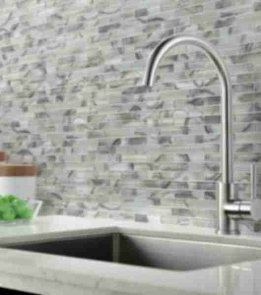 Linear glass mosaic tiled backsplash behind a kitchen sink with a silver faucet.