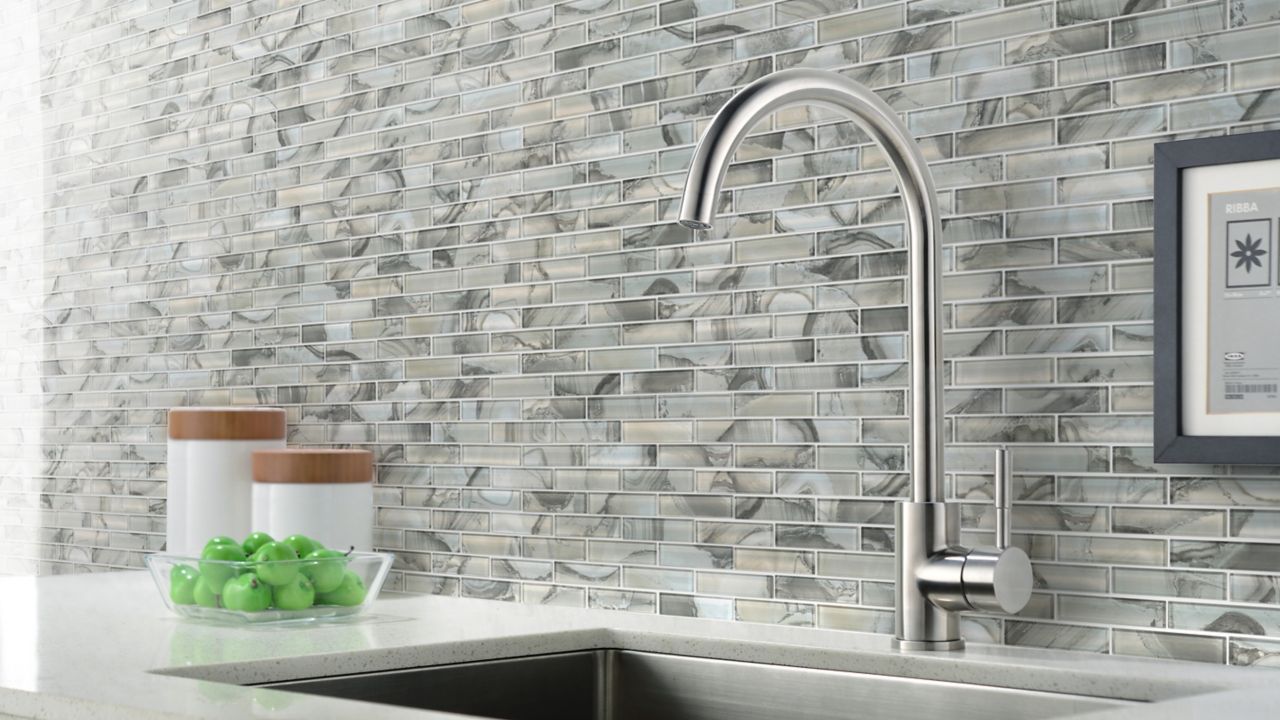 A sink surrounded by white countertops, gray cabinets underneath, and a glass tile backsplash that is a combination of brown, blue and beige.