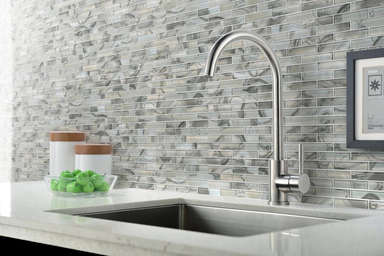 Linear glass mosaic tiled backsplash behind a kitchen sink with a silver faucet.