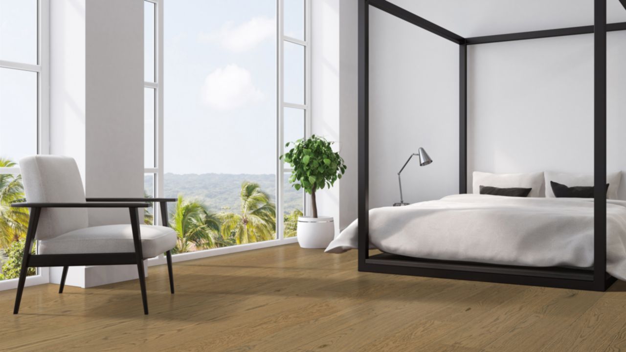 A bedroom white white bed, white chair, and light brown engineered hardwood. Room has large windows overlooking palm trees.