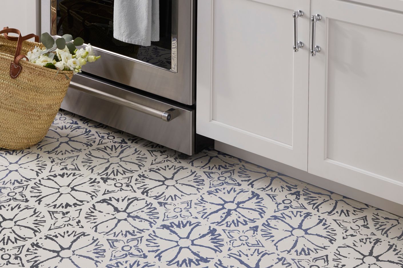 Tile kitchen area featuring a pattern tile on the floor a pattern tile as an accent behind the stove top field tile is a subway tile in white with a natural edge countertops are a slate grey cabinetry white and appliances are stainless. Coordinating tile profiles are used to divide accent from the field tile in the backsplash.