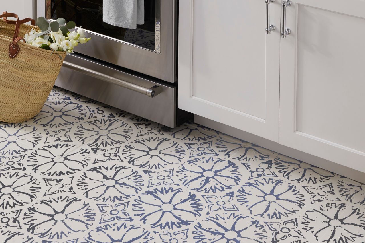 Tile kitchen area featuring a pattern tile on the floor a pattern tile as an accent behind the stove top field tile is a subway tile in white with a natural edge countertops are a slate grey cabinetry white and appliances are stainless. Coordinating tile profiles are used to divide accent from the field tile in the backsplash.