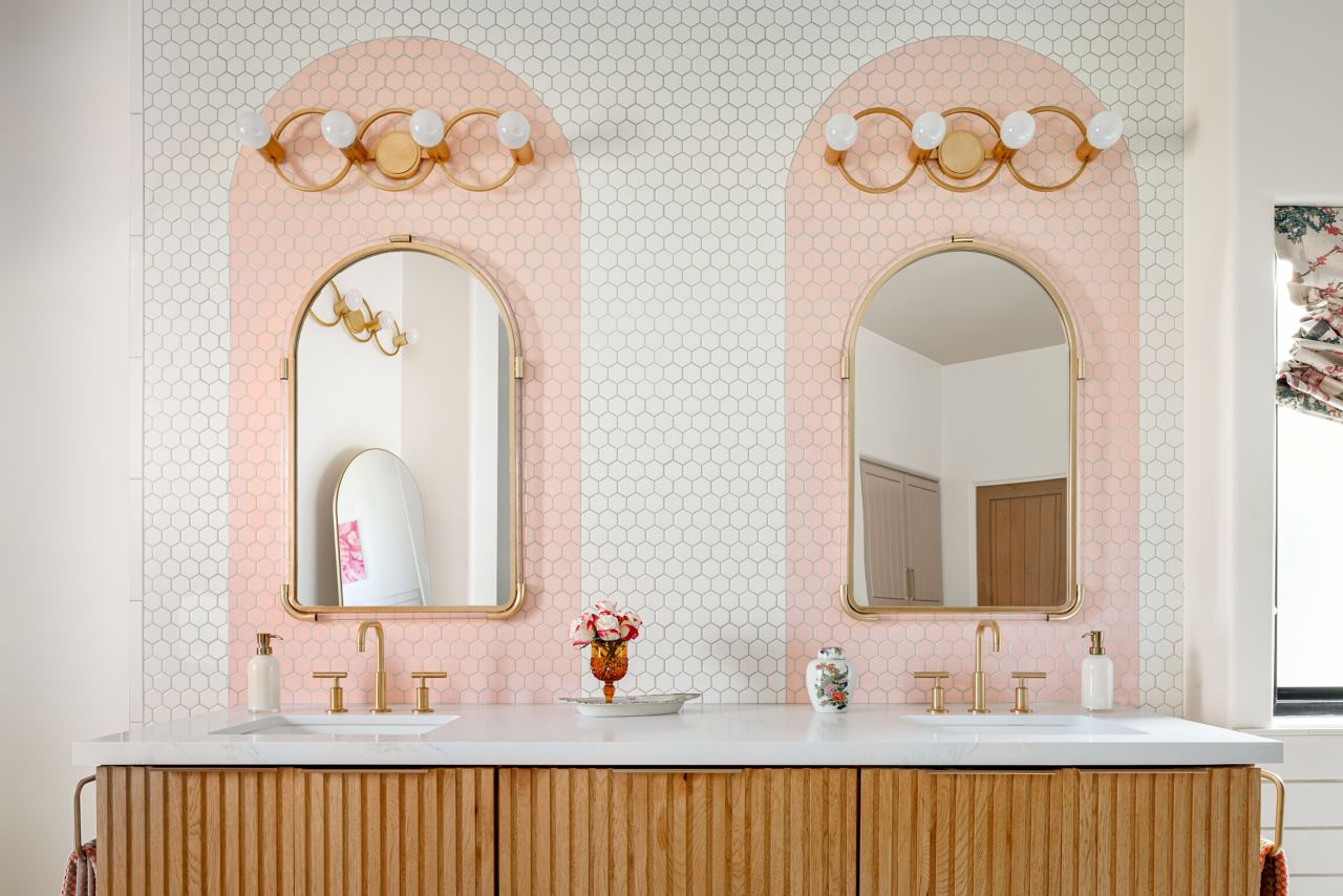 Double sink vanity with mirrors, gold light fixtures and white with pink arch tile