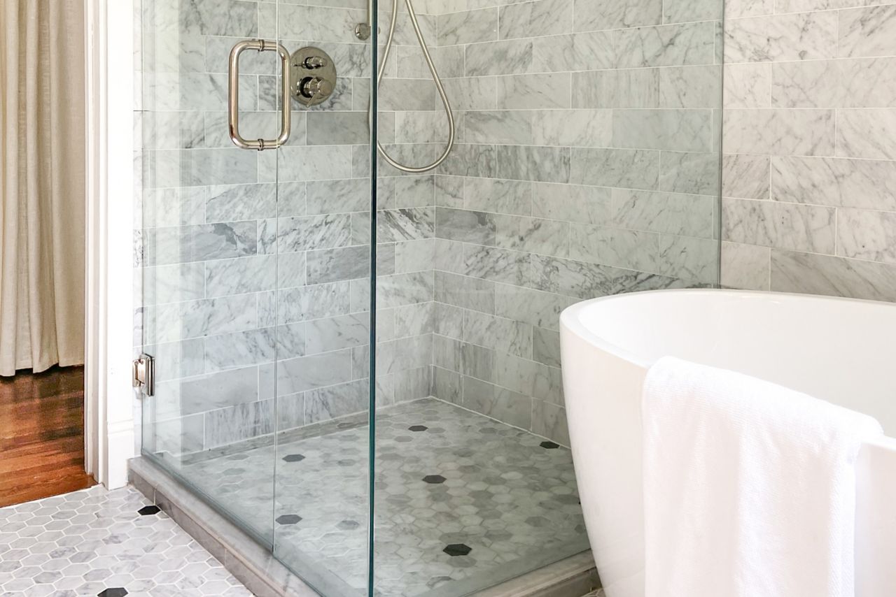 Glass-enclosed shower with white and grey marble tile wall. The floor is grey and white marble hexagons with a pattern of black marble hexagon polka dots.