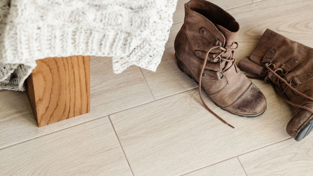 
        A pair of worn brown leather ankle boots with laces sits on a light-colored wood-look tile floor next to a piece of furniture draped with a cable-knit blanket.