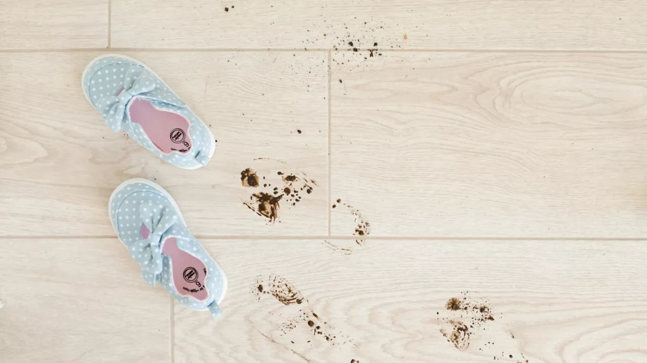 An overhead view of a light wood-look tile floor with a pair of light blue, polka-dotted children's shoes next to a trail of small, muddy footprints and dirt splatters.