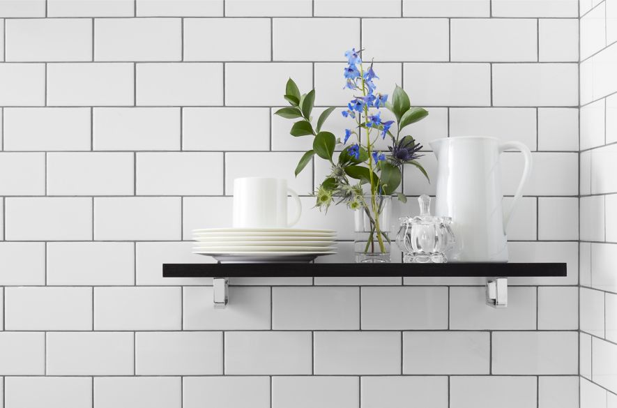 White subway tile with dark grout on wall with black shelf holding miscellaneous tableware and greenery in a vase.