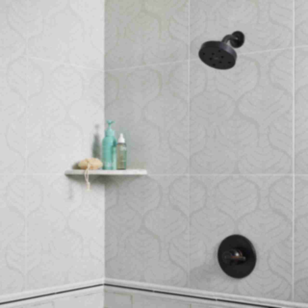 A bathroom shower design combines patterned tile on the upper shower walls, with subway tile on the lower shower walls. Trim pieces are layered to create an attractive transition between the two surface materials, and tie in the color of the shower fixtures. A small corner shelf on the upper portion of the wall adds functional storage.