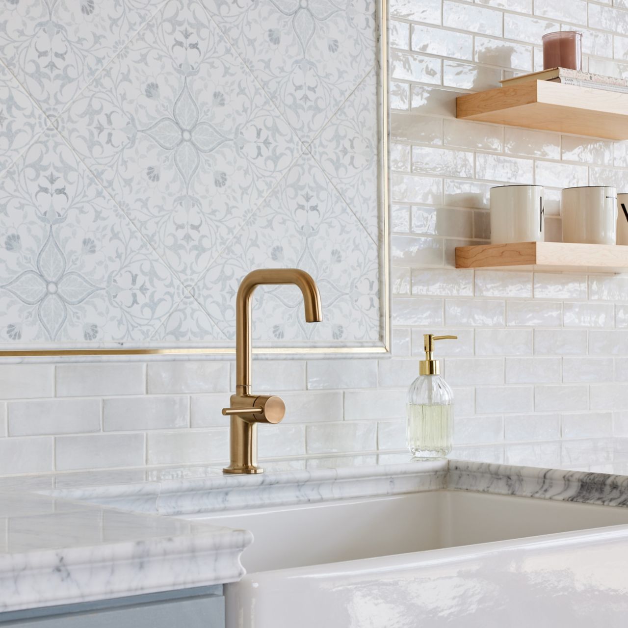 The wall behind a kitchen sink is completely tiled to create a large backsplash area. Patterned porcelain tile is framed with gold pencil trim pieces to create a decorative focal point directly above the sink, and glossy white subway tile is used for the surrounding wall. Floating shelves in a light wood tone flank either side of the patterned tile accent.