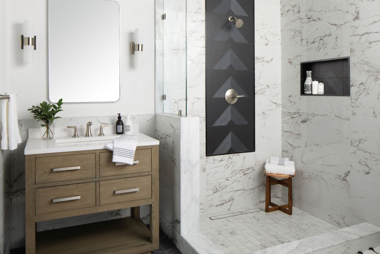 Bathroom area with walk-in shower with a porcelain bodied- faux marble looking tile on walls with black picture frame accent around shower fixtures and in the back of recessed niche shower pan is of matching tile and a smaller caliber like wall tile - wainscoting tile to match shower wall tile  surrounds rest of room small shower stool sits in shower area bathroom vanity is of natural wood fixtures of our of a brushed silver metal mirror and sconces match.