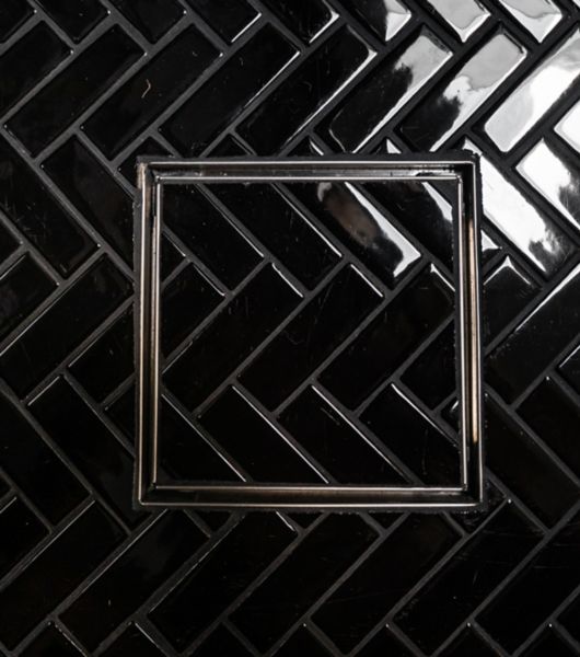 The floor of a shower is covered in glossy black porcelain mosaic tile featuring a herringbone arrangement. The shower drain is also covered in the same tile, providing visual continuity.