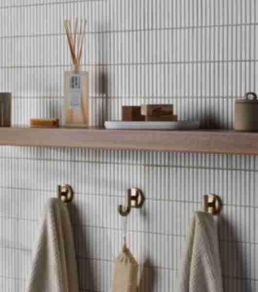 A bathroom wall features open shelving and gold-tone towel hooks over white porcelain tile that resembles small white bars stacked vertically.
