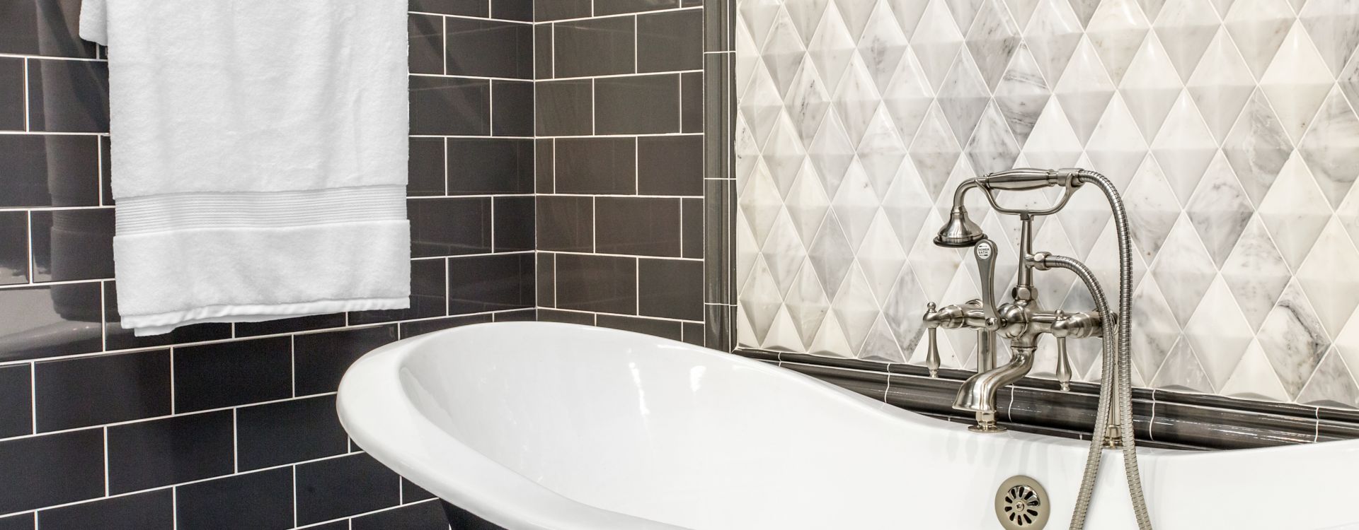 Bathroom area with soaker tub subway tile used on walls with matching trim and baseboard decorative picture frame behind tub is a dimensional carrara marble in a diamond shape Grays whites and blacks are the colors in the space tile mimics colors and tub and metal fixtures are a soft silver appearance.