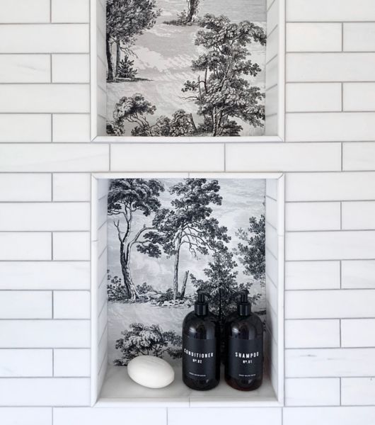 A shower wall, covered in white subway tile with dark grout, features two recessed storage niches. The niches are tiled with a wallpaper-look tile in a traditional black and white landscape motif.