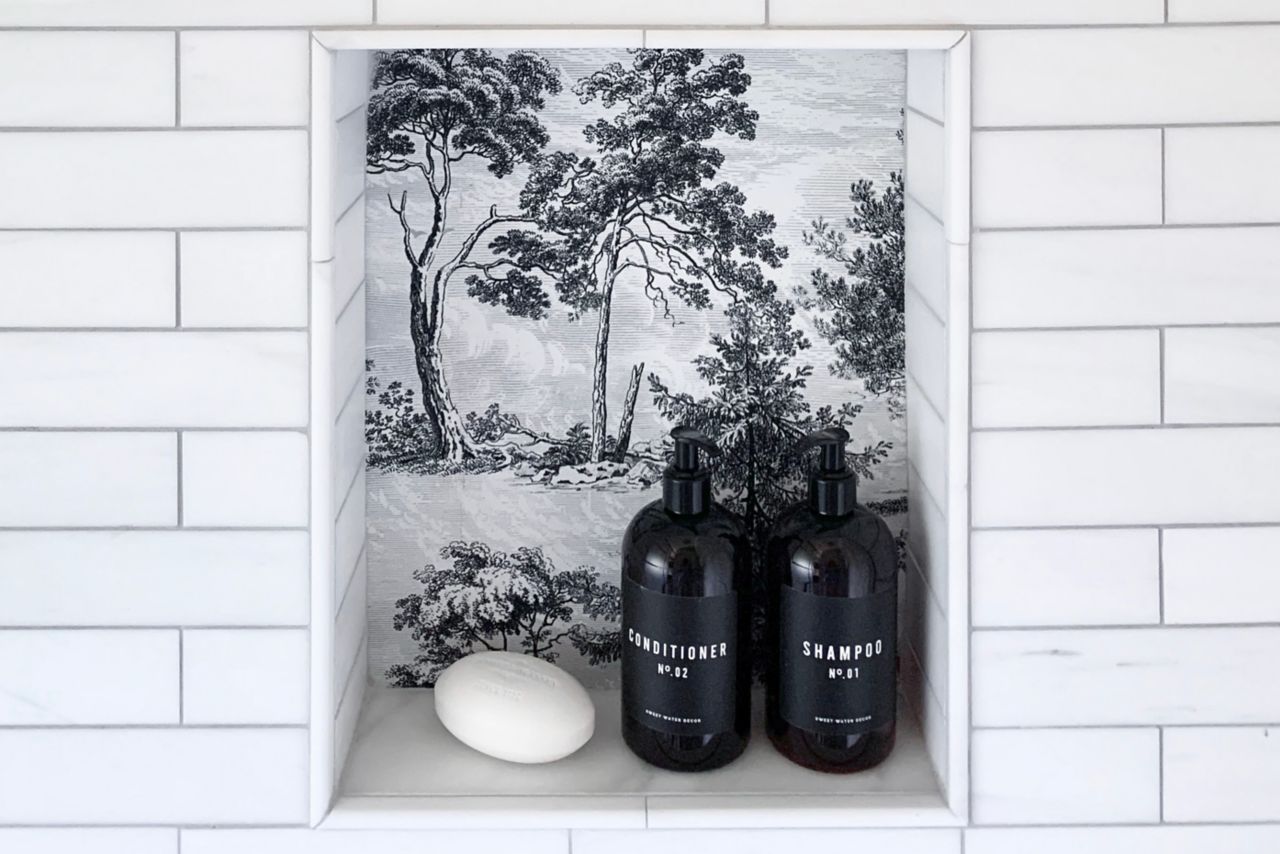 A shower wall, covered in white subway tile with dark grout, features two recessed storage niches. The niches are tiled with a wallpaper-look tile in a traditional black and white landscape motif.