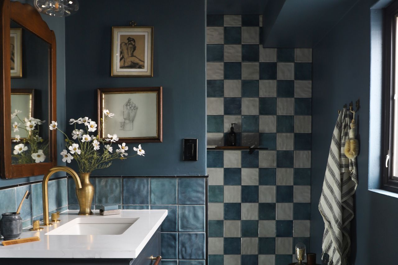 Bathroom with dark blue vanity and wall paint, and dark blue tiles on walls. Past the vanity is a walk-in shower with a floor-to-ceiling wall of dark blue and off-white tile arranged in a checkerboard pattern. The floor is covered in a mosaic of dark blue tiles with occasional small accents of white tile.